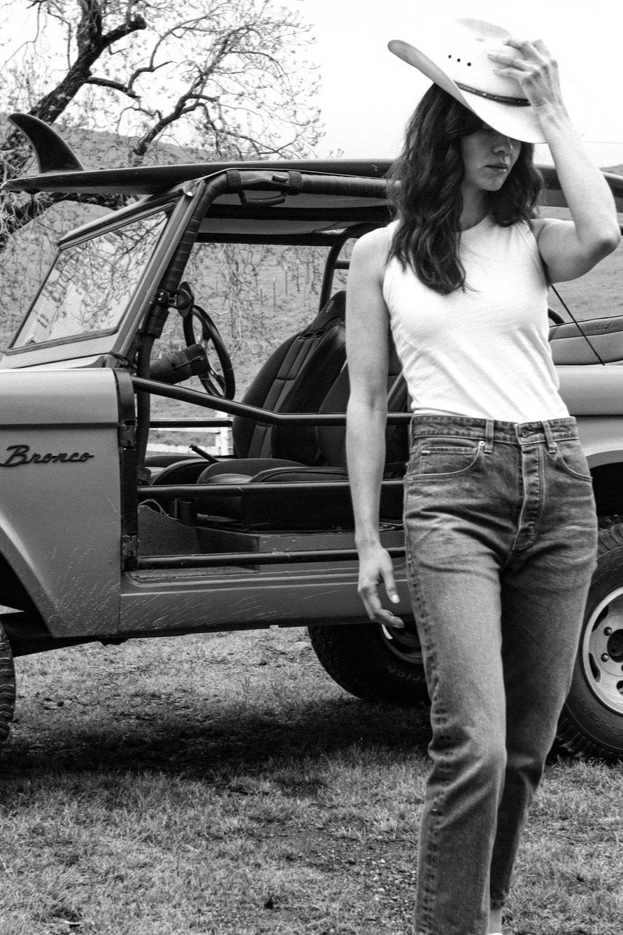 Woman in a sleeveless Reyes tank and jeans wearing a cowboy hat, standing beside a vintage Bronco.