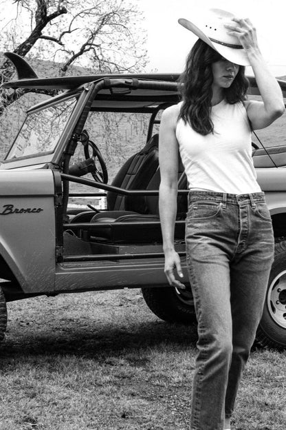 Woman in a sleeveless Reyes tank and jeans wearing a cowboy hat, standing beside a vintage Bronco.
