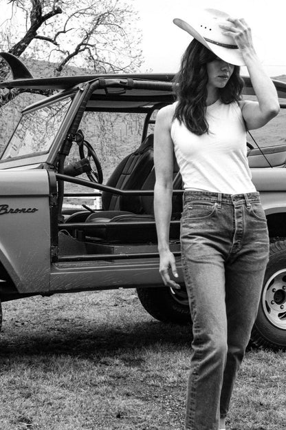Model in white Reyes tank and jeans, wearing a cowboy hat beside a vintage Bronco.