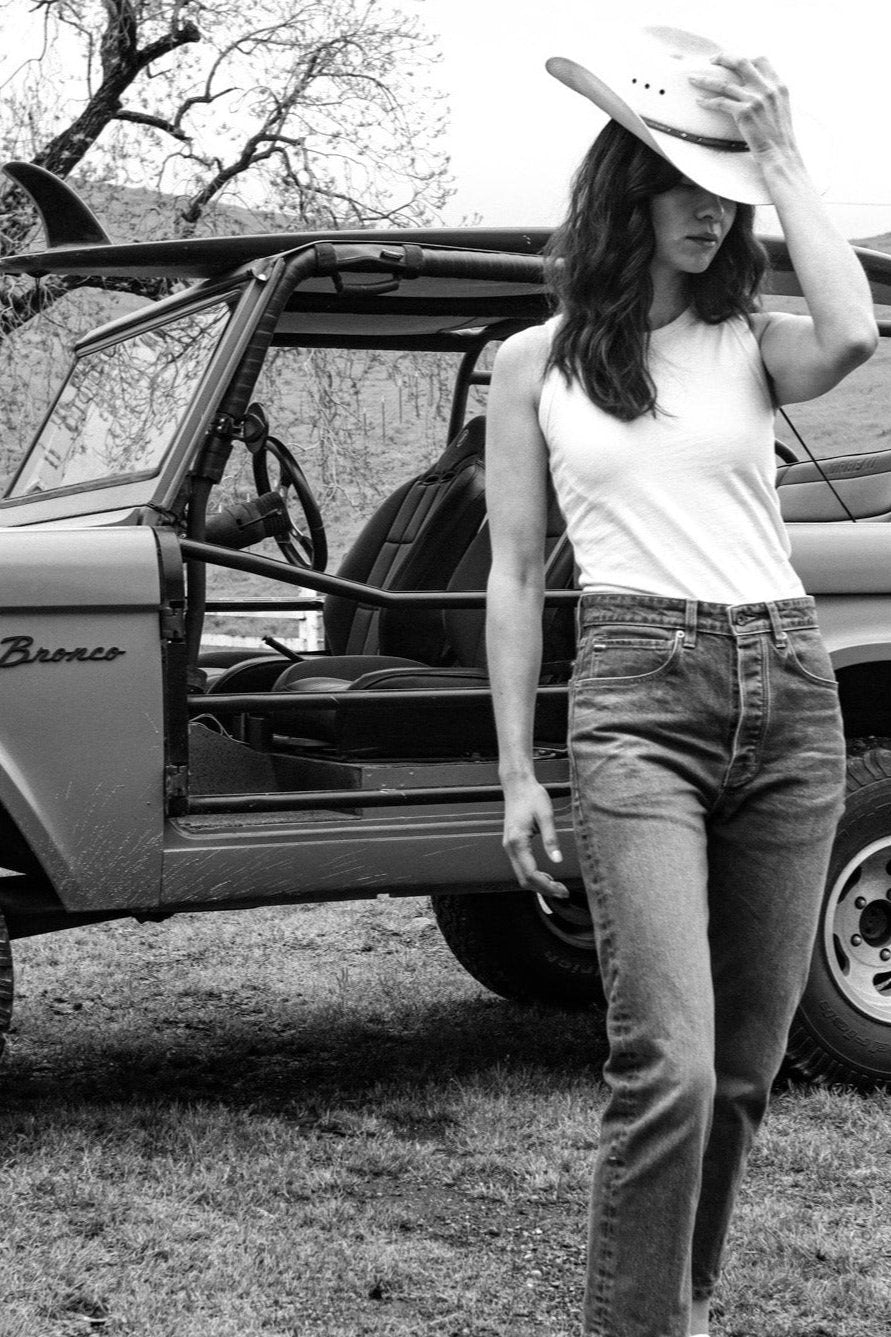 Woman in white Reyes cotton tank and jeans wearing a cowboy hat beside a vintage Bronco.