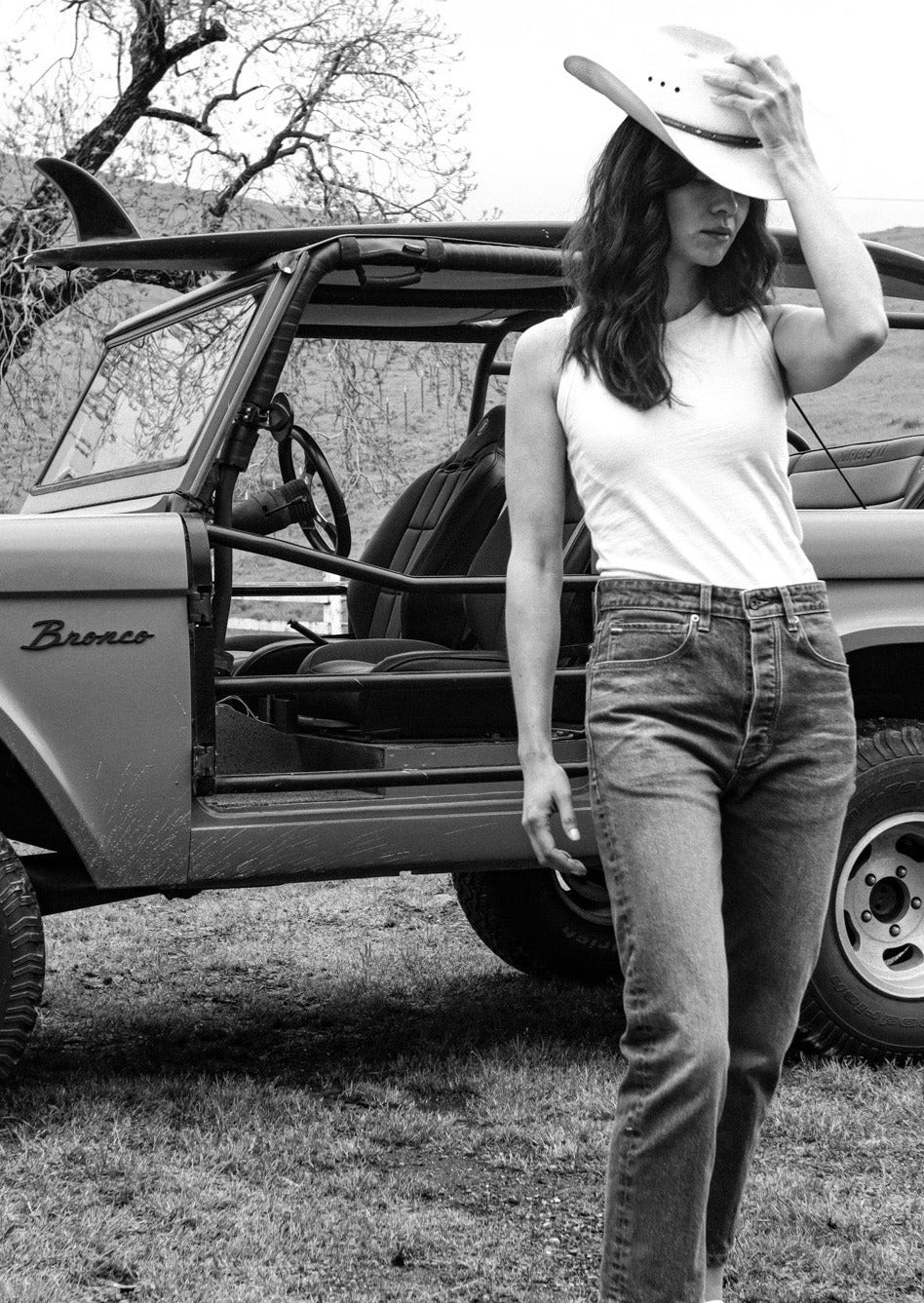 Woman in a white Bolinas cotton tank and jeans wearing a cowboy hat beside a vintage Bronco.