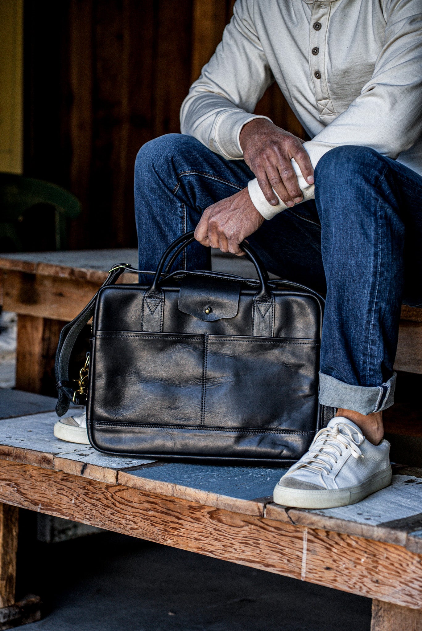 Black Horween leather briefcase with brass hardware, held by a seated person in denim.