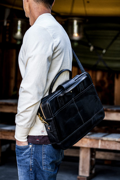 Black Horween leather briefcase with brass zipper carried on the shoulder outdoors.