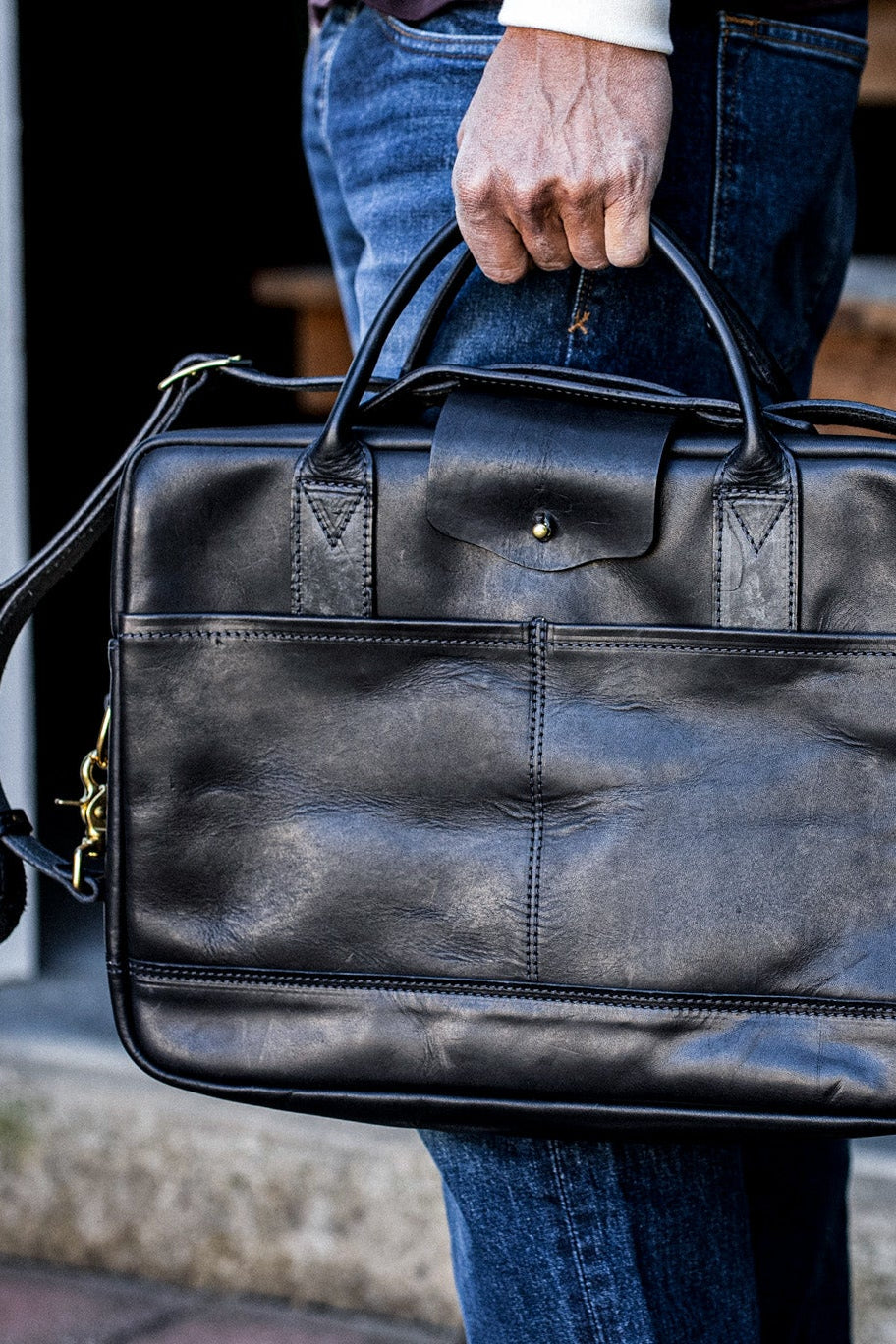 Black Horween leather briefcase with brass hardware, carried by a person in denim jeans.