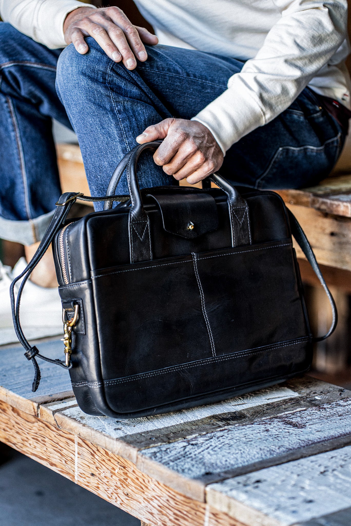 Black Horween leather briefcase with brass zipper, held on a wooden bench by a seated person.