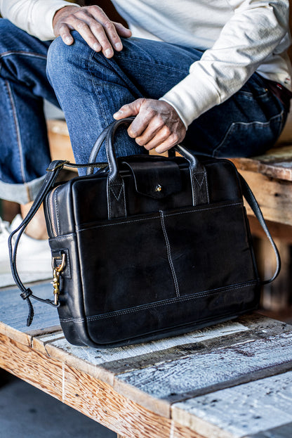 Black Horween leather briefcase with brass zipper, held on a wooden bench by a seated person.
