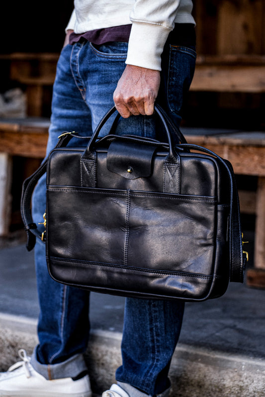 Black Horween leather briefcase with brass zipper, carried by hand over dark denim jeans.