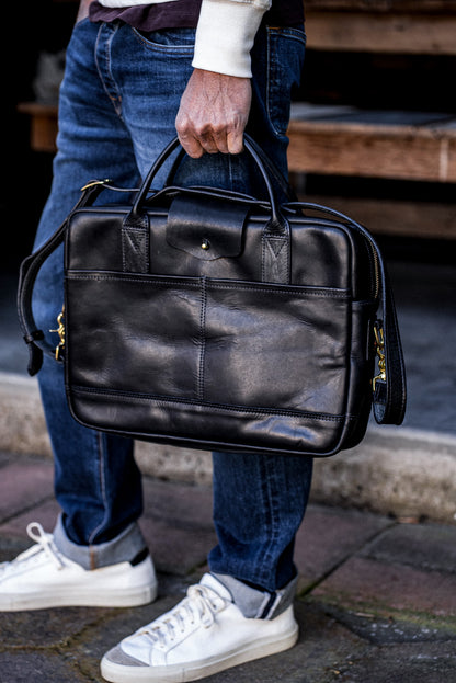Black Horween leather briefcase with brass zipper, held by hand against denim jeans.