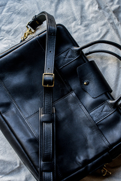 Black Horween leather briefcase with brass buckle and adjustable shoulder strap on linen.