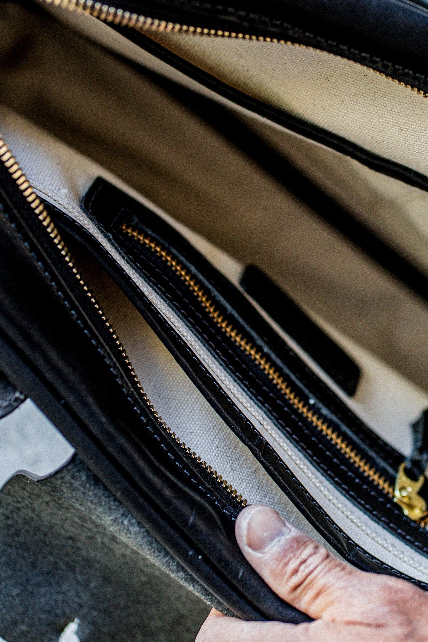 Hand opening a black Horween leather brief, revealing natural canvas lining and brass zipper pockets.