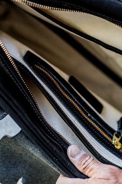 Hand opening a black Horween leather brief, revealing natural canvas lining and brass zipper pockets.