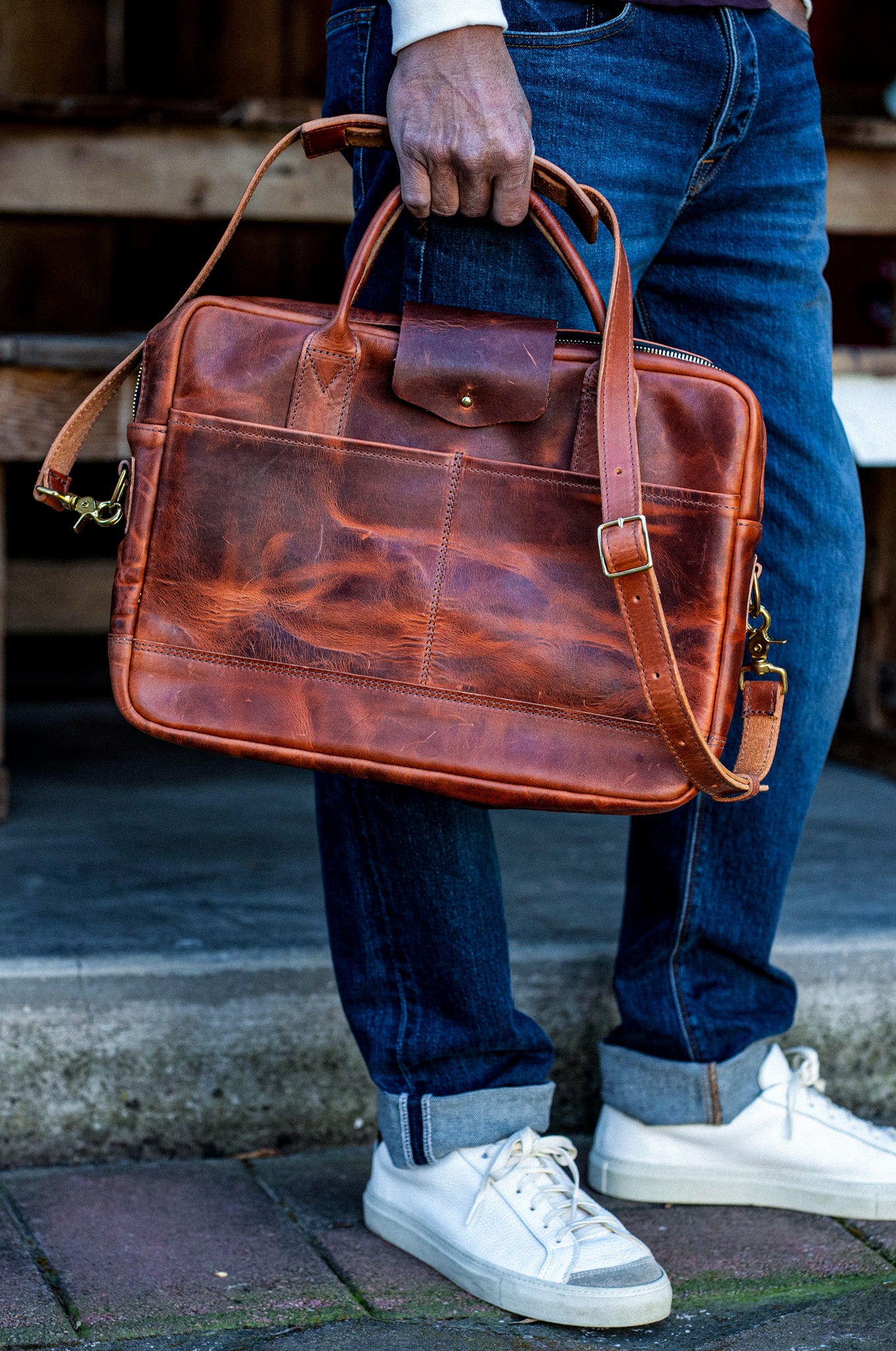Person holding a tan Horween leather briefcase with brass hardware and shoulder strap.