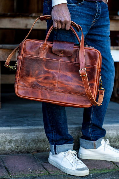 Person holding a tan Horween leather briefcase with brass hardware and shoulder strap.