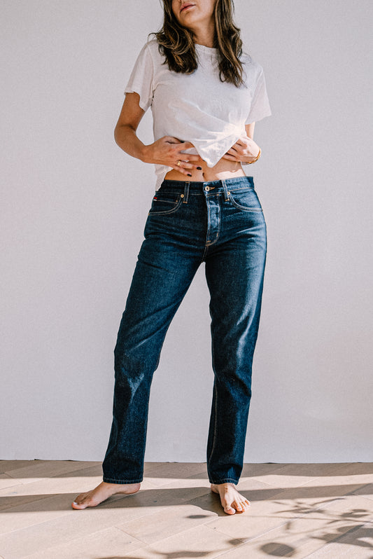 Model in a white tee and dark ocean wash mid-rise slim fit American made jeans, standing barefoot on wood floor.