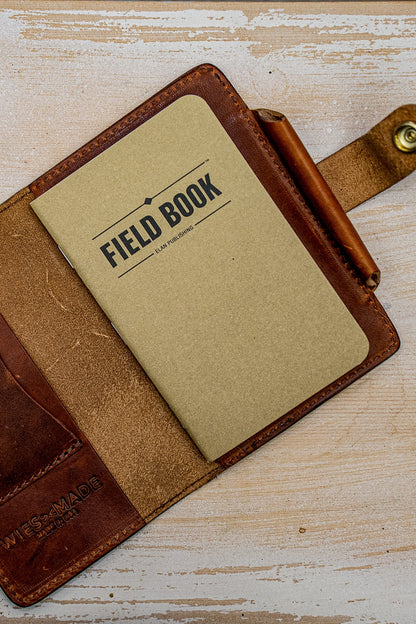 Open tan Horween leather field notes cover holding a notebook and pen with brass snap.