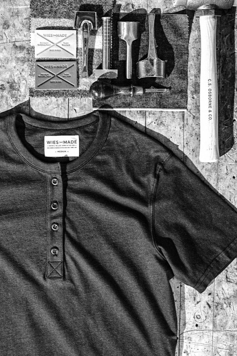 Basalt gray four-button Reyes Henley shirt laid flat beside leather labels and metal craft tools.