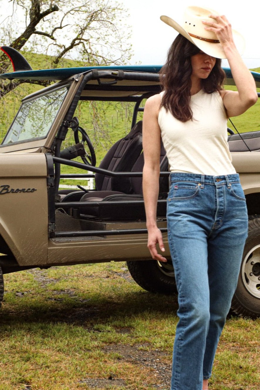 Woman in natural cotton Bolinas tank and blue jeans standing beside a vintage Bronco in a grassy field.
