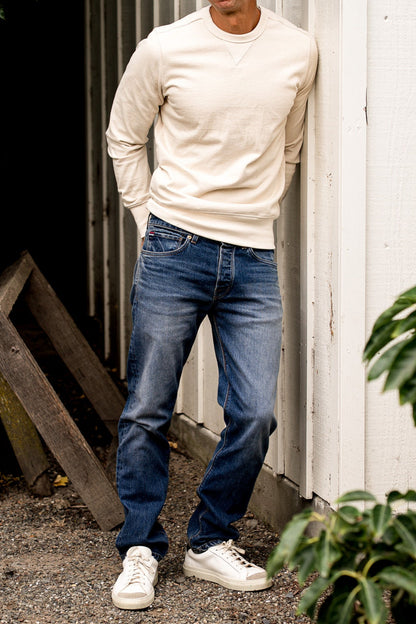 Man wearing men's denim jeans made in USA with cream sweatshirt and white sneakers, leaning against a white wall