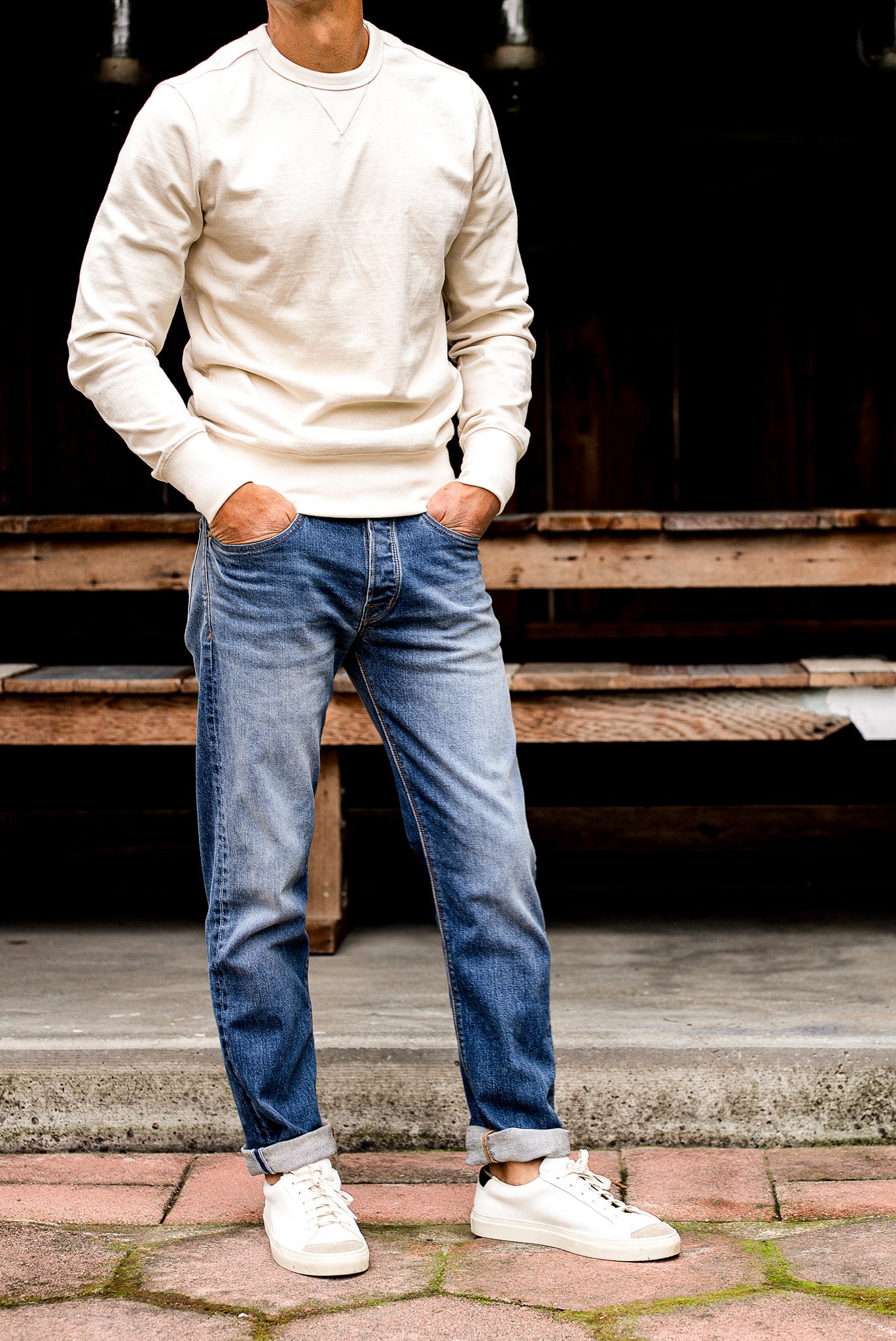 Man wearing men's jeans made in USA with cuffed straight leg fit, cream sweater, and white sneakers