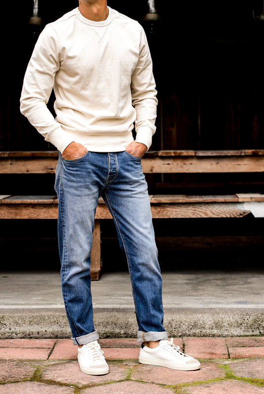 Man wearing men's jeans made in USA with cuffed straight leg fit, cream sweater, and white sneakers