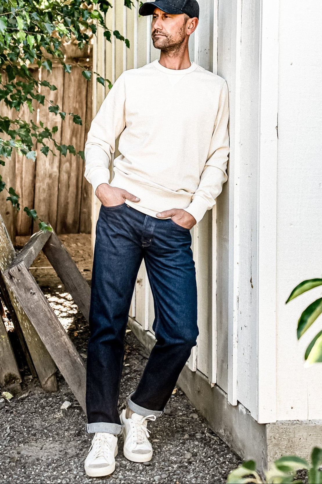 Man in cream sweatshirt and cap wearing dark ocean wash loose straight American made jeans with cuffed hems.