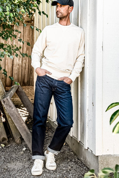 Man in cream sweatshirt and cap wearing dark ocean wash loose straight American made jeans with cuffed hems.