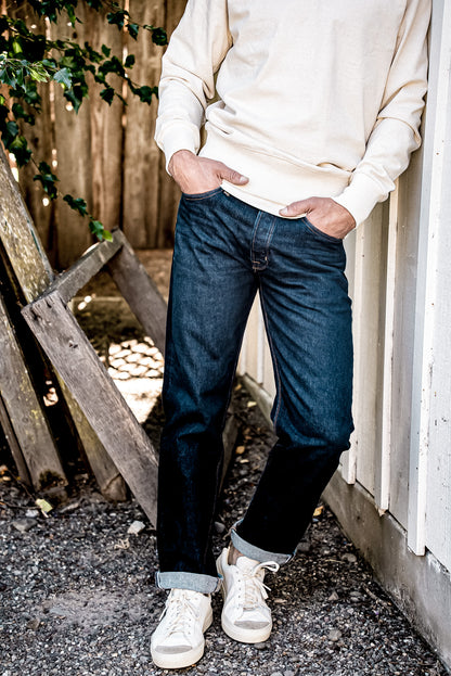 Man in cream sweatshirt and cuffed ocean wash loose straight American made denim jeans with white sneakers.