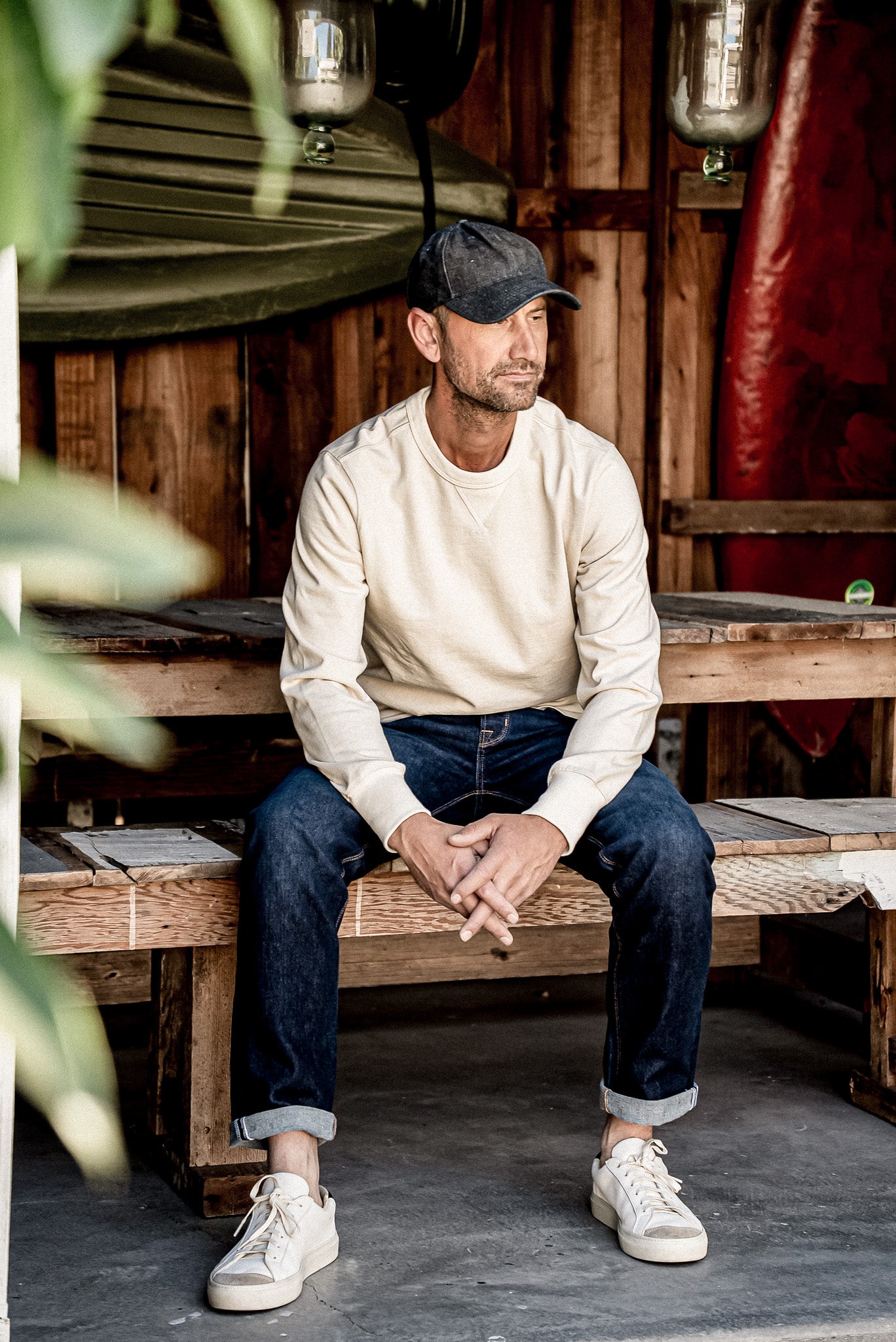 Man in cuffed dark wash American made jeans and cream sweatshirt seated on a wooden bench.