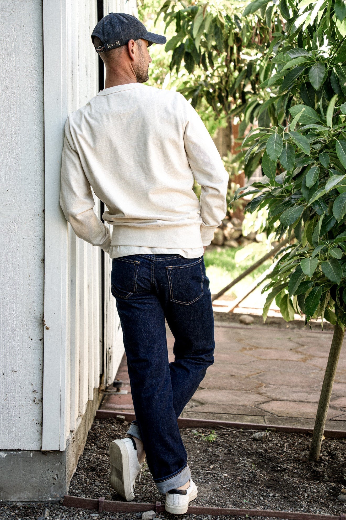 Back view of man in dark ocean wash loose straight cut jeans with cuffed hems, cream sweatshirt, and cap.