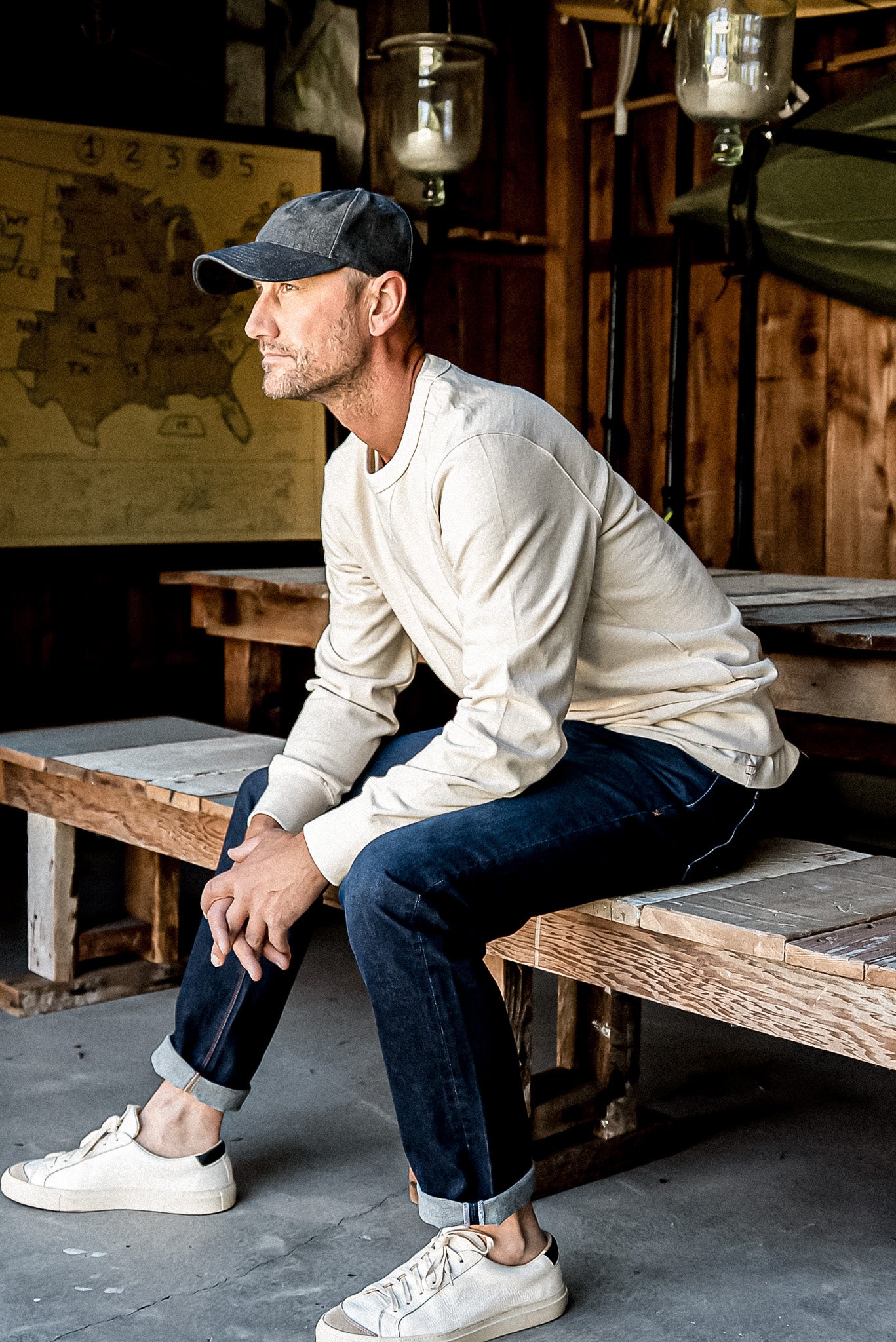 Man in cuffed dark wash American made jeans and cream sweatshirt sitting on a wooden workshop bench.