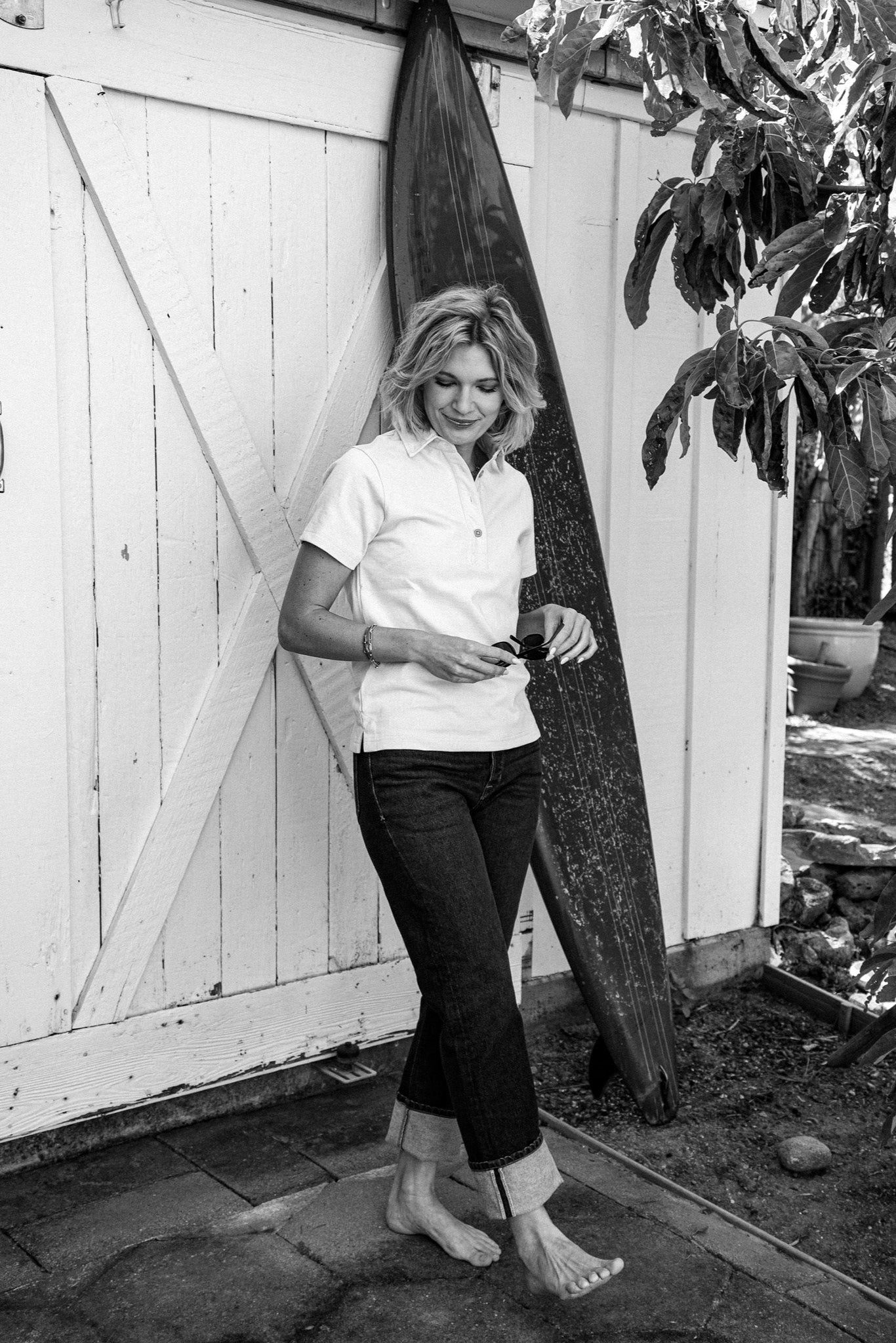 Model in cuffed Jackie wide-leg jeans and white polo leaning against a surfboard by a barn door.