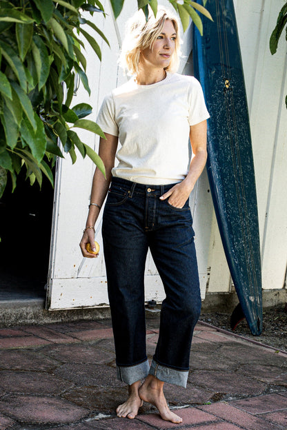Woman in a cream tee and cuffed wide-leg American-made jeans standing barefoot beside a surfboard.