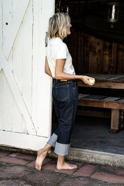 Side view of cuffed dark indigo wide-leg jeans paired with a cream tee, worn barefoot beside a white barn door.