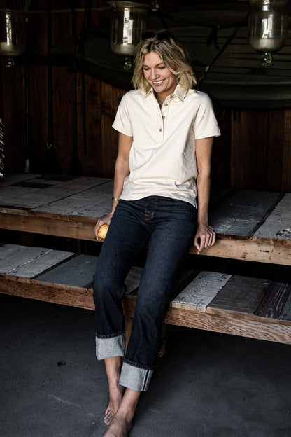 Woman in a cream polo and cuffed dark ocean wash wide-leg jeans seated on a weathered wooden bench.