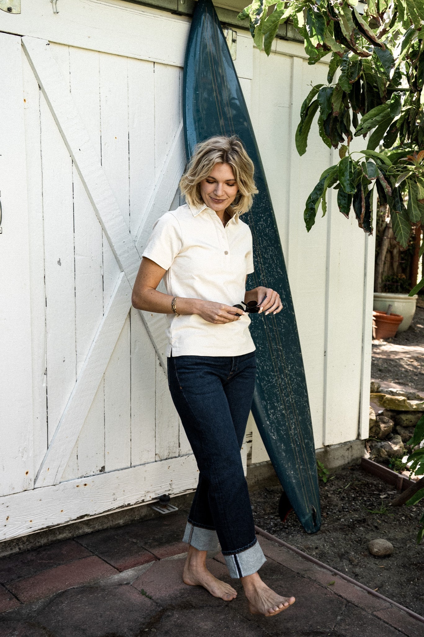 Person in cuffed ocean wash wide-leg jeans leaning on a barn door with a surfboard.