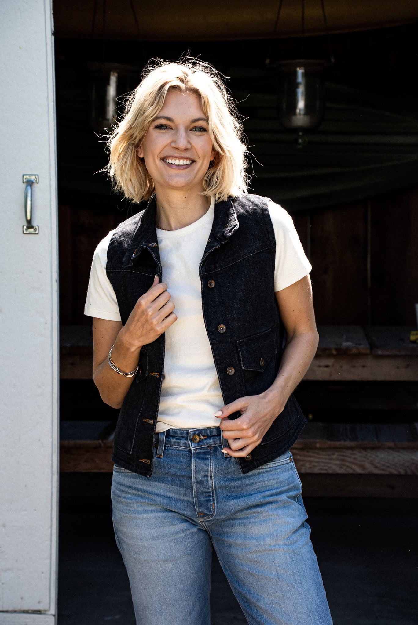 Woman wearing a black broken twill denim vest over a cream tee and light wash jeans.