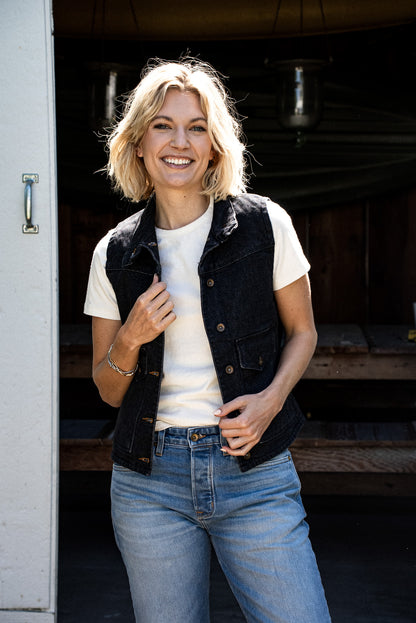 Woman wearing a black broken twill denim vest over a cream tee and light wash jeans.