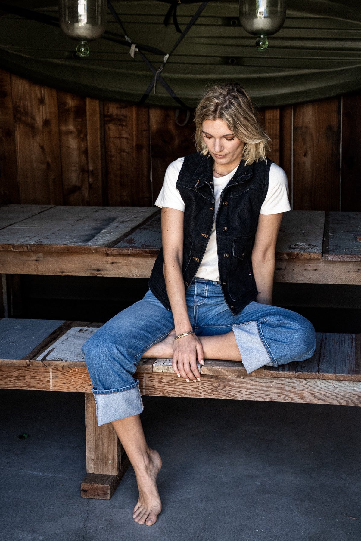 Person in black broken twill denim vest and cuffed jeans on a rustic wooden bench.