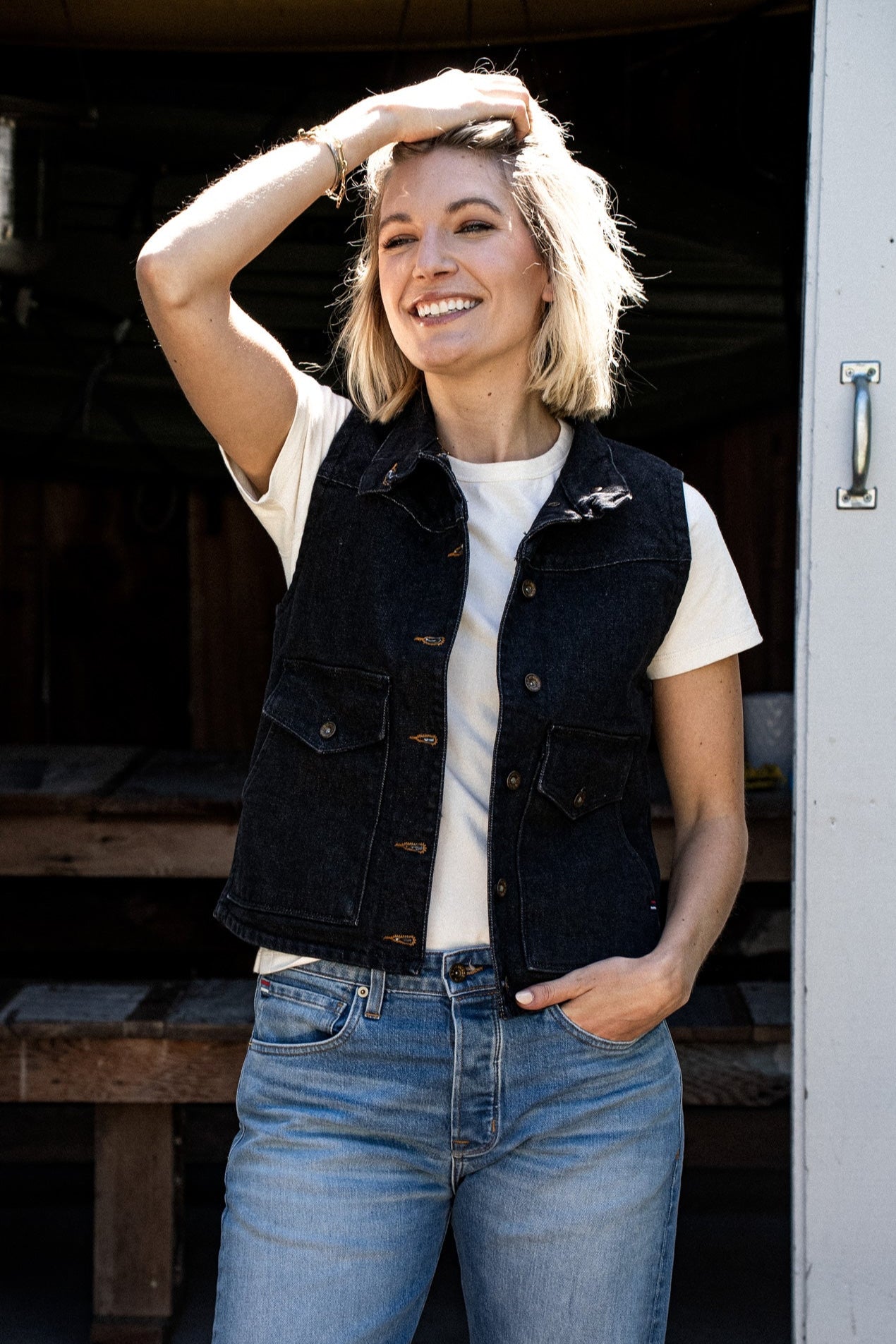 Woman smiling in a black broken twill denim vest layered over a white tee with light wash jeans.