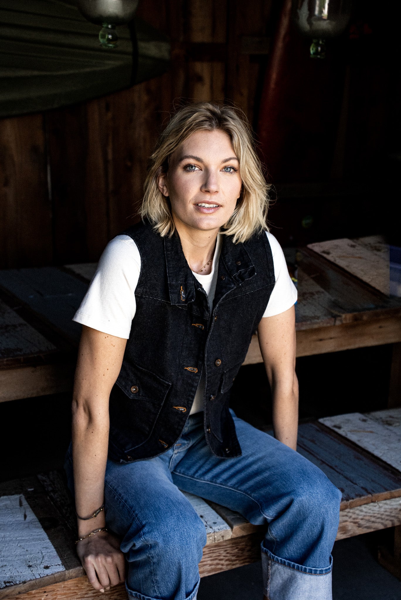 Person wearing black broken twill denim Jamie vest over a white tee, seated on rustic wood.