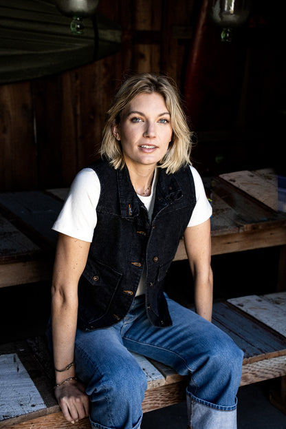 Person wearing black broken twill denim Jamie vest over a white tee, seated on rustic wood.