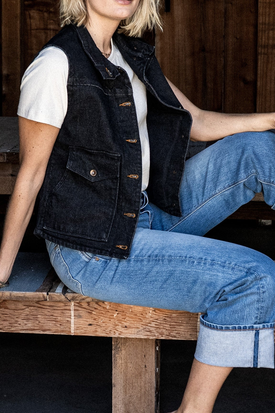 Woman seated on a wooden bench wearing a black broken twill denim vest over a white tee with cuffed light-wash jeans.
