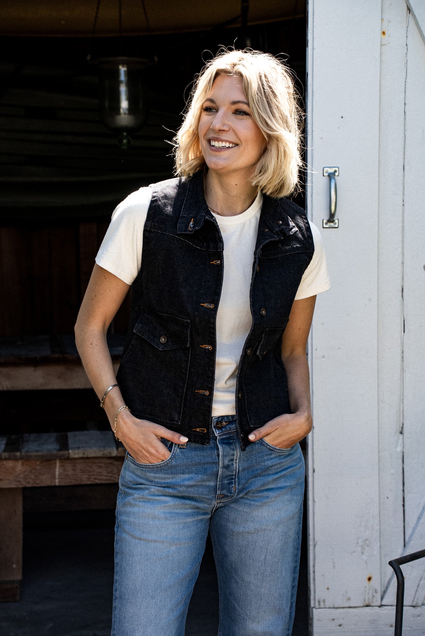 Woman wearing black broken twill denim vest over a cream tee with light-wash jeans.