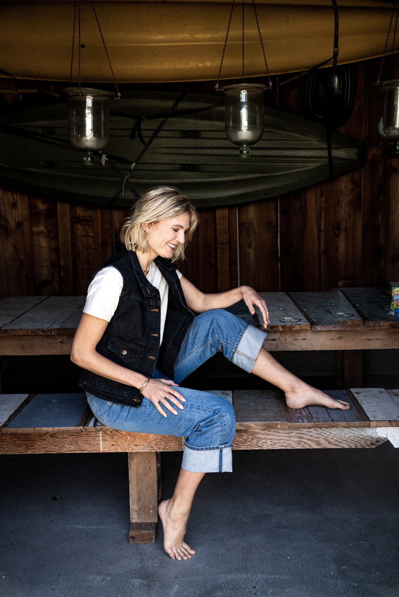 Woman in a black broken twill denim vest and cuffed blue jeans sitting on a wooden bench under hanging canoes.