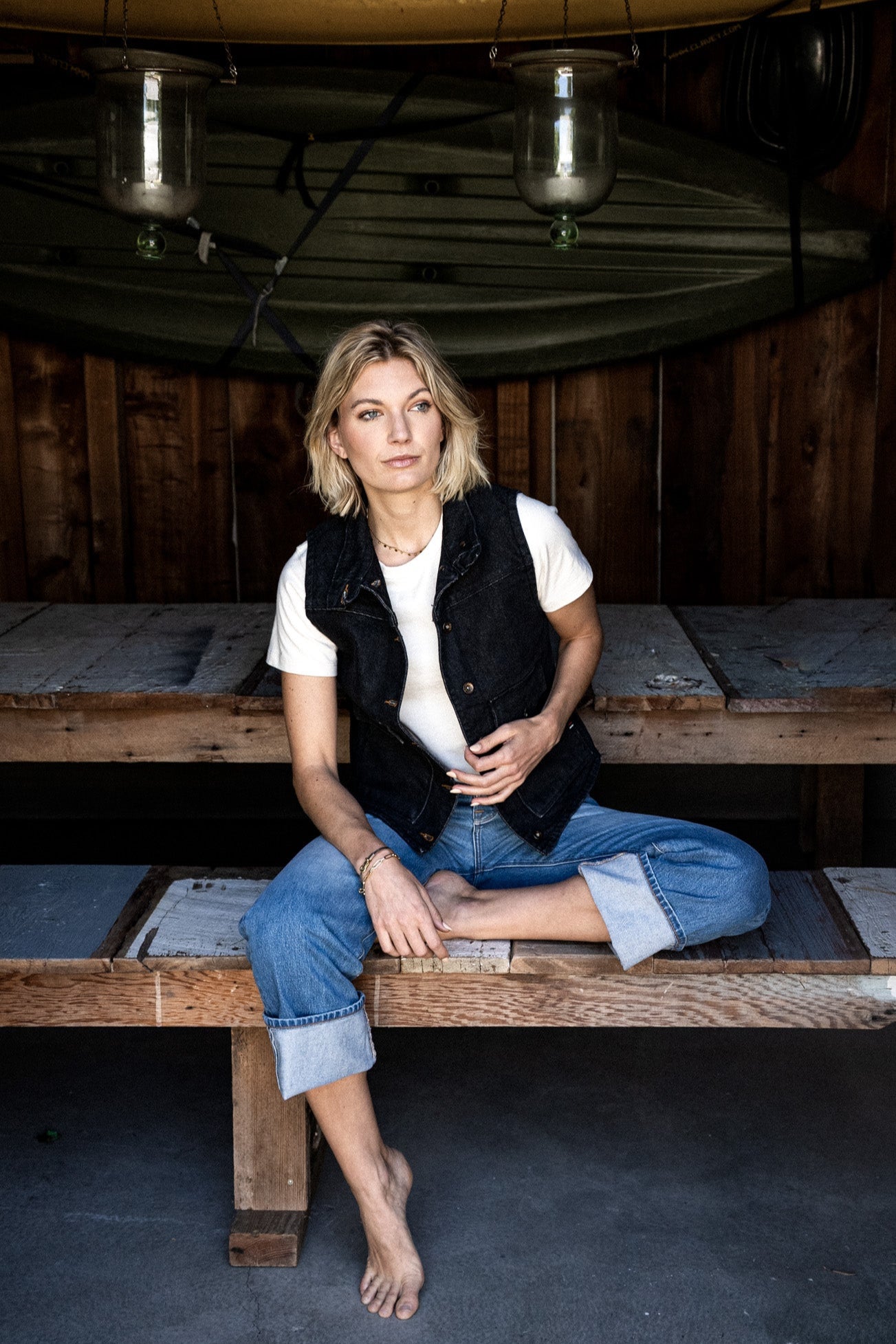 Model wears black broken twill denim Jamie vest over a white tee, seated on a wooden bench.