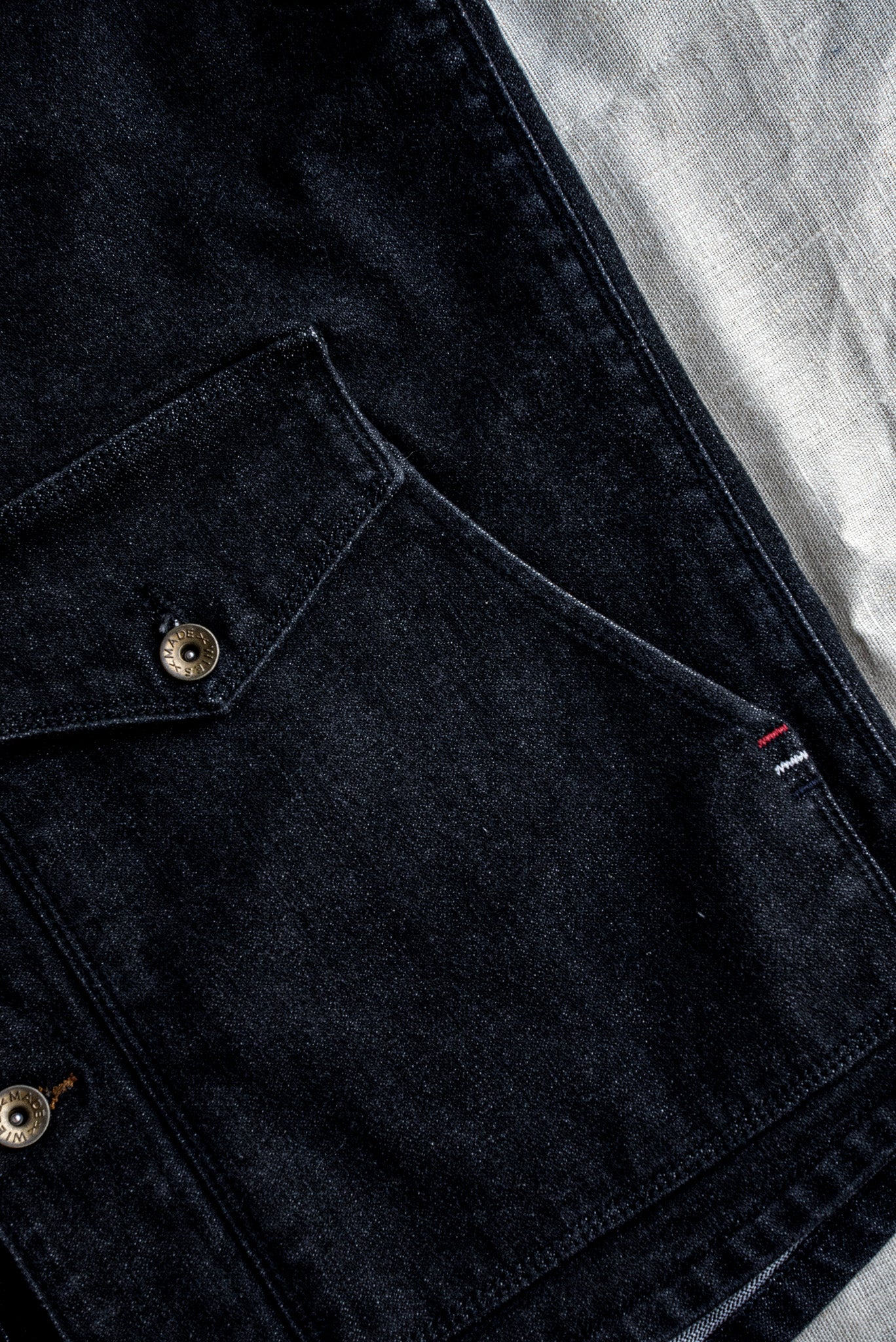 Close-up of black broken twill denim with brass Wies buttons and red-white-blue bartack stitching.