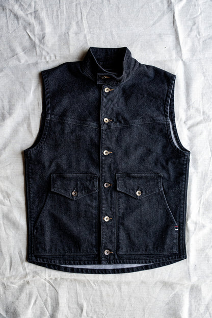 Black broken twill denim vest laid flat, showing brass buttons and chest pockets.