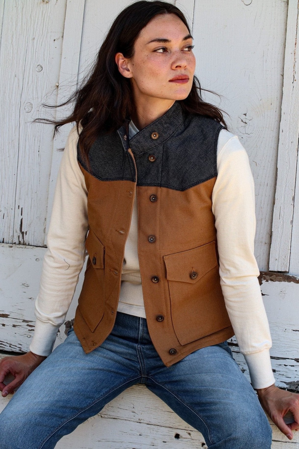 Woman wearing tan canvas Jamie vest with denim yoke, brass buttons, seated against white barn wood.