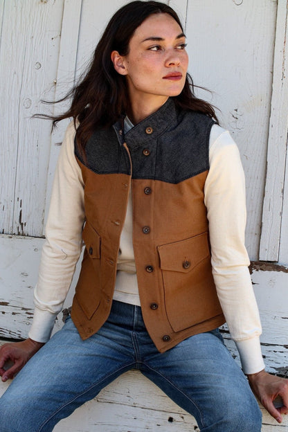 Woman wearing tan canvas Jamie vest with denim yoke, brass buttons, seated against white barn wood.
