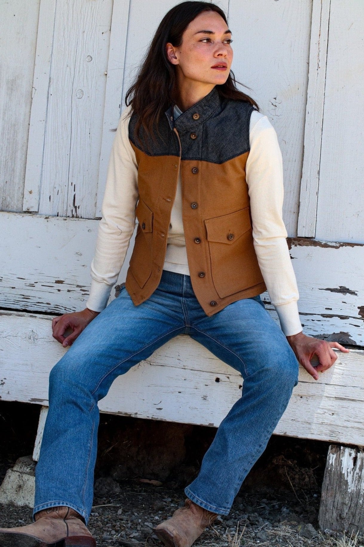 Woman in a tan canvas and denim-yoke Jamie vest over a cream sweater and blue jeans, seated on a weathered white bench.
